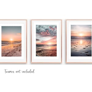 May include: Three vertical wall art prints featuring sunset beach scenes with pink and orange hues, displayed in rose gold frames with white matting. The text "Frames not included" is printed in black script below the three framed images.