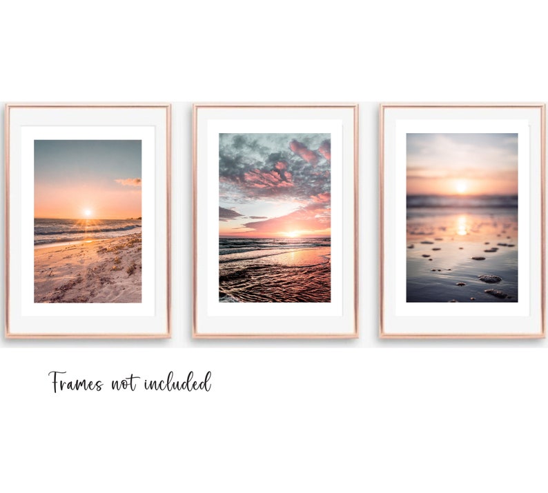 May include: Three vertical wall art prints featuring sunset beach scenes with pink and orange hues, displayed in rose gold frames with white matting. The text "Frames not included" is printed in black script below the three framed images.
