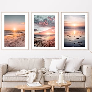 May include: Three framed vertical wall art prints featuring beach sunset scenes. The left print shows a sandy shore at sunrise, the center print displays a vibrant pink and blue cloudy sky over ocean waves, and the right print captures a blurred sunset.