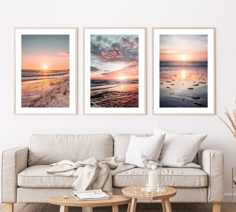 May include: Three framed vertical wall art prints featuring beach sunset scenes. The left print shows a sandy shore at sunrise, the center print displays a vibrant pink and blue cloudy sky over ocean waves, and the right print captures a blurred sunset.