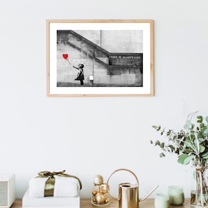 May include: Black and white photograph of a street art mural depicting a young girl reaching for a red heart-shaped balloon. The text "There is always hope" is written on the wall.