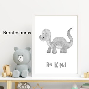 Puede incluir: Una impresi&oacute;n enmarcada con una ilustraci&oacute;n gris y blanca de un dinosaurio Brontosaurus. El texto "Be Kind" est&aacute; debajo del dinosaurio. La impresi&oacute;n est&aacute; en un estante blanco con un oso de peluche gris y otros art&iacute;culos decorativos.