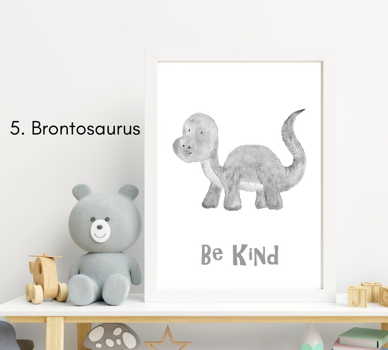 Puede incluir: Una impresi&oacute;n enmarcada con una ilustraci&oacute;n gris y blanca de un dinosaurio Brontosaurus. El texto "Be Kind" est&aacute; debajo del dinosaurio. La impresi&oacute;n est&aacute; en un estante blanco con un oso de peluche gris y otros art&iacute;culos decorativos.