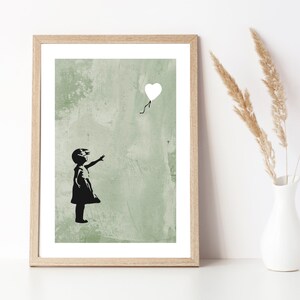 May include: A black and white print of a girl reaching for a white heart-shaped balloon. The background is a green textured wall.