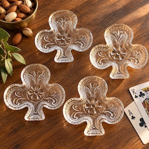May include: Four clear glass dishes shaped like the club suit from a deck of cards. Each dish features a central embossed flower motif surrounded by intricate scrollwork and a textured stippled pattern along the edges of the clover shape.