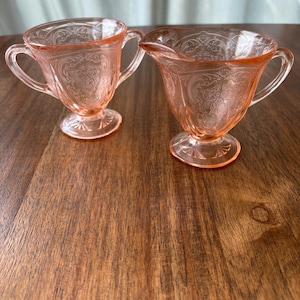 May include: A pair of pink depression glass vessels, consisting of a two-handled sugar bowl and a creamer with a pouring spout. Both pieces feature a pedestal base and an intricate embossed floral pattern across the translucent pink surface.