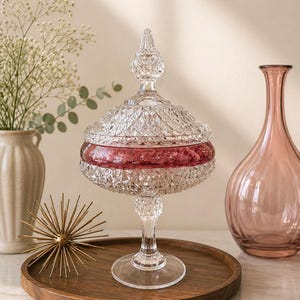 May include: A clear glass pedestal candy dish with a diamond-cut pattern and a removable lid. The center of the bowl features a wide, horizontal band of deep pink glass. The dish stands on a circular base and is topped with a pointed finial.