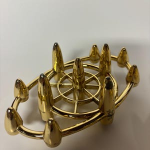 May include: A gold-colored metal candle holder with a circular design. The holder features a central wheel-like structure and multiple conical spikes around the perimeter. The overall design is symmetrical and ornate.