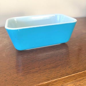May include: A rectangular, turquoise-colored Pyrex baking dish. The dish has a light blue interior and white trim. The dish is sitting on a wooden surface.