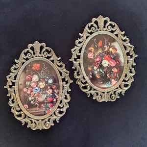 May include: Two oval wall plaques featuring floral still life prints encased in ornate, metallic gold-toned frames with intricate scrollwork and leaf-like detailing. Each print depicts a colorful bouquet of flowers arranged in a dark vase.