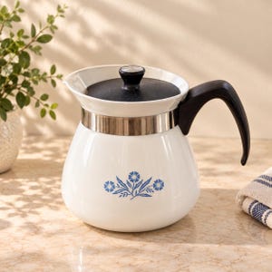 May include: A white ceramic teapot with a black lid and a black handle. The body of the teapot features a blue floral pattern centered on the front. A silver-colored metal band encircles the upper portion of the teapot just below the lid.