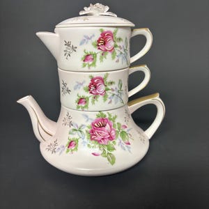 May include: A stacked, three-piece porcelain tea set. The set is white with pink roses and green leaves. The teapot has a spout and handle, and the lid is topped with a decorative rose. The cups have gold trim.