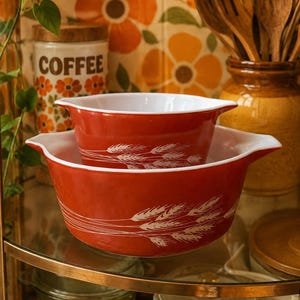 May include: Two nested orange glass mixing bowls with white interiors and white wheat stalk patterns on the exterior. Each bowl features two side handles and a smooth, rounded rim. The bowls are stacked one inside the other on a glass shelf.