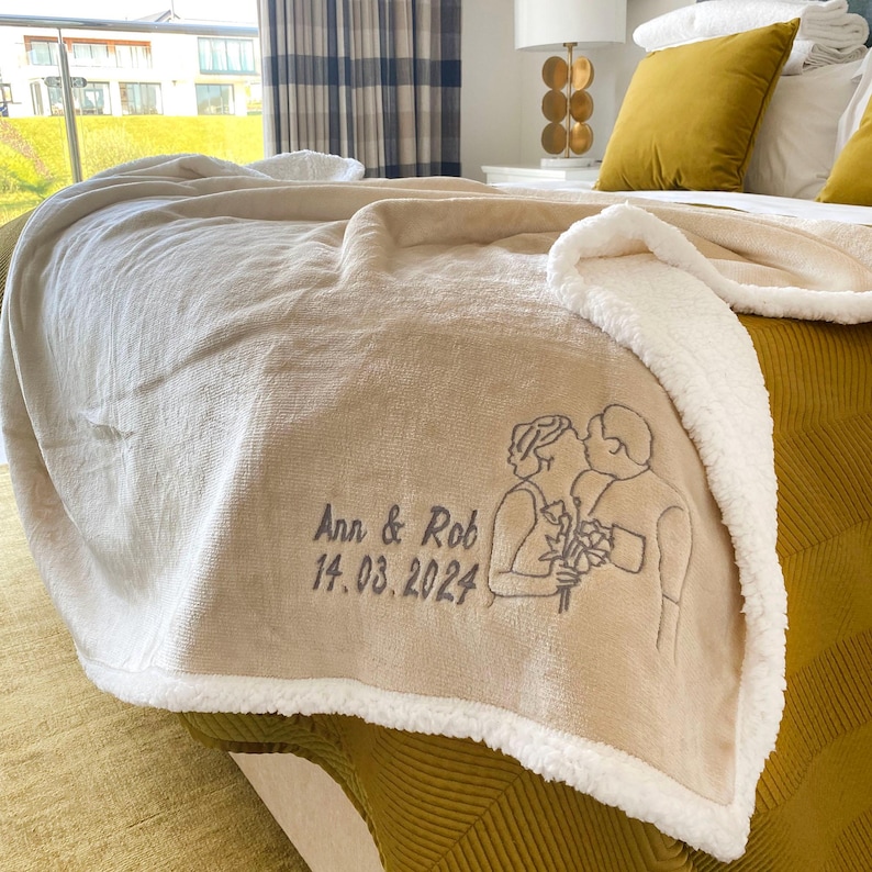 Pode incluir: Uma manta de arremesso bege com forro de sherpa branco. A manta apresenta um design bordado de um casal com o texto "Ann & Rob 14.03.2024".