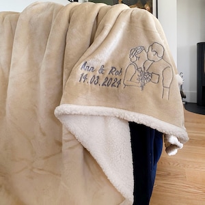 Pode incluir: Uma manta de l&atilde; de carneiro bege com forro de sherpa branco. A manta tem um desenho bordado de um casal com o texto "Ann & Rob 14.03.2024".