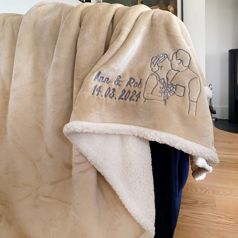 Pode incluir: Uma manta de l&atilde; de carneiro bege com forro de sherpa branco. A manta tem um desenho bordado de um casal com o texto "Ann & Rob 14.03.2024".