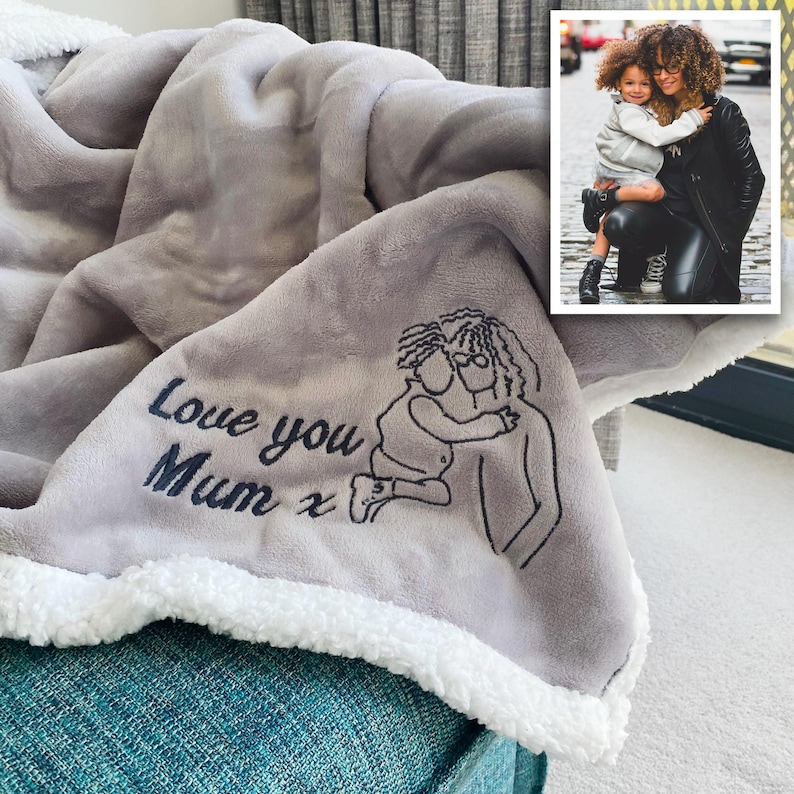 Pode incluir: Cobertor de l&atilde; cinza com uma borda de sherpa branca. O cobertor tem um desenho bordado de duas pessoas se abra&ccedil;ando com o texto "Love you Mum x".