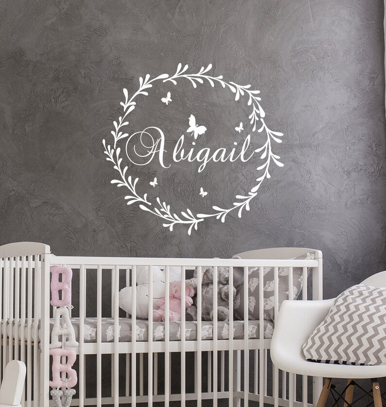 May include: White vinyl wall decal with a floral wreath and the name Abigail in a cursive font. The wreath is decorated with small butterflies. The decal is displayed on a gray wall above a white crib.