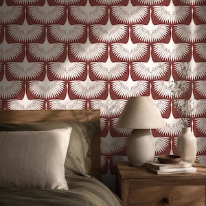 May include: Wallpaper featuring a repeating pattern of white stylised birds with spread wings against a deep red background. The design is displayed behind a wooden bed frame with olive green bedding, a beige lamp, and a ceramic vase with dried stems.