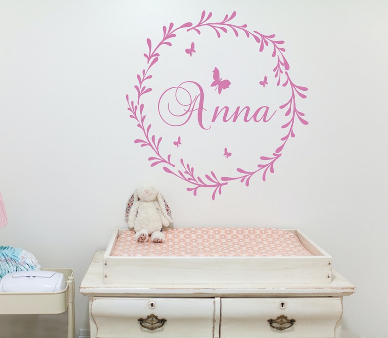 May include: Pink floral wreath wall decal with the name "Anna" in a cursive font and several small butterfly silhouettes.
