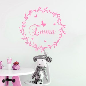 May include: Pink wall decal with a floral wreath and the name "Emma" in a cursive font.  The wreath is decorated with small pink butterflies.