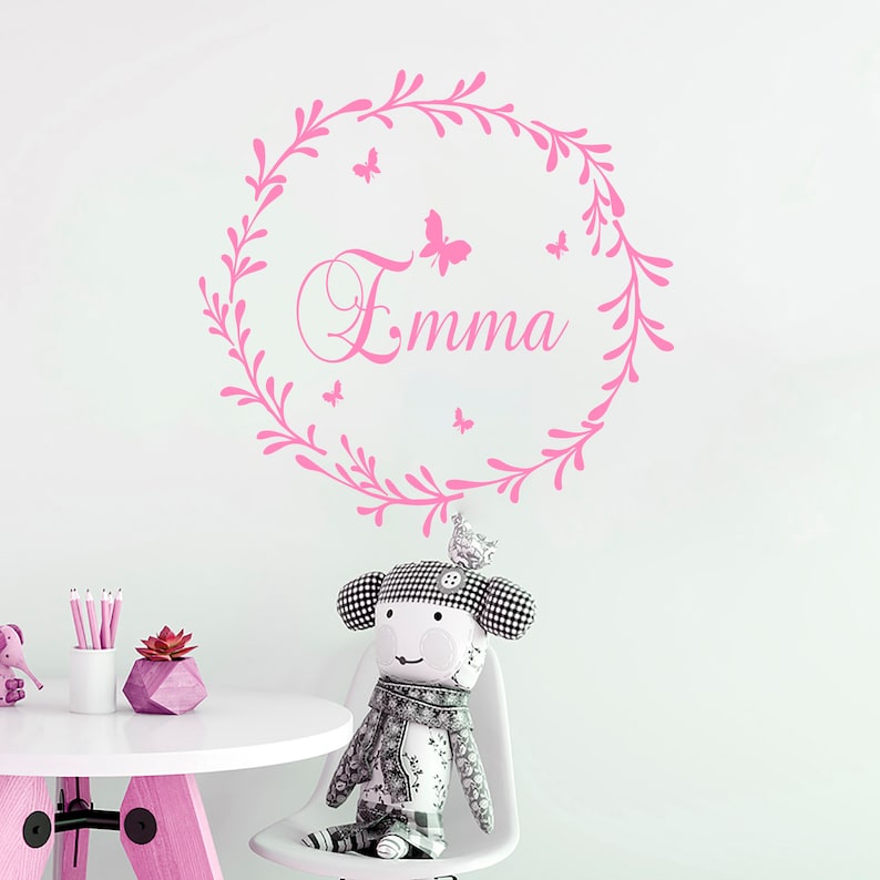 May include: Pink wall decal with a floral wreath and the name "Emma" in a cursive font.  The wreath is decorated with small pink butterflies.