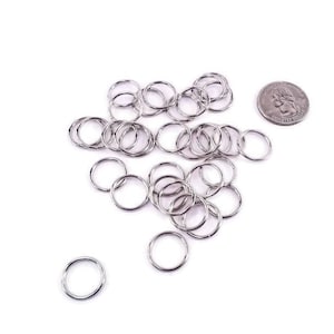May include: A pile of silver jump rings, approximately the size of a US quarter, on a white background.