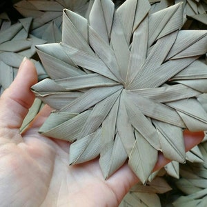 May include: A hand holds a woven star-shaped ornament made from dried palm leaves. The ornament is light brown and has a natural, rustic look.