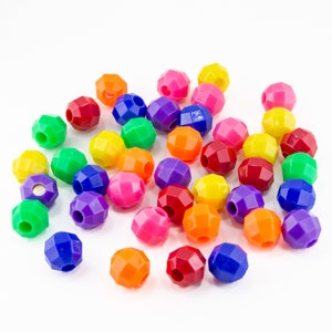 May include: A collection of colorful, faceted beads in various shades including red, orange, yellow, green, blue, purple, and pink. Each bead has a hole for stringing. The beads are scattered on a white surface.