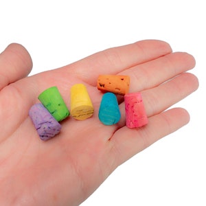 May include: A hand holding six small, colorful cork stoppers. The stoppers are painted in shades of purple, green, yellow, blue, orange, and pink.