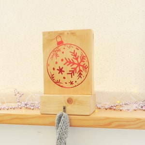 Puede incluir: Un soporte de calcetines de Navidad de madera con un diseño de adorno rojo. El soporte tiene un gancho para colgar un calcetín.