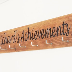 Könnte beinhalten: Eine Holztafel mit dem Text "Richard's Achievements" und Metallhaken zum Aufhängen von Medaillen oder anderen Gegenständen.