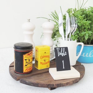 May include: A wooden table number holder with a black chalkboard sign that says "Table 1". The holder is on a wooden tray with a pepper shaker, a mustard jar, and a white jug with a plant and cutlery.