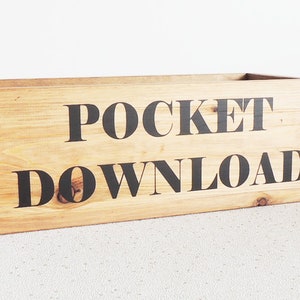 Puede incluir: Una caja de madera con las palabras "POCKET DOWNLOAD" impresas en la parte delantera en letras negras.