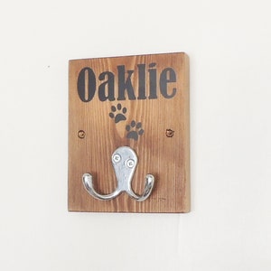 May include: A wooden dog leash hook with the name "Oaklie" and paw prints painted on it. The hook is attached to a wall.