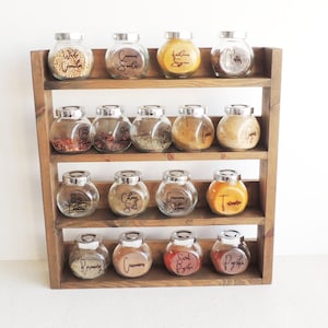 May include: A wooden spice rack with three shelves holds 15 glass jars with silver lids. Each jar has a handwritten label with the name of a spice, such as "White Pepper", "Cinnamon Sticks", and "Turmeric".