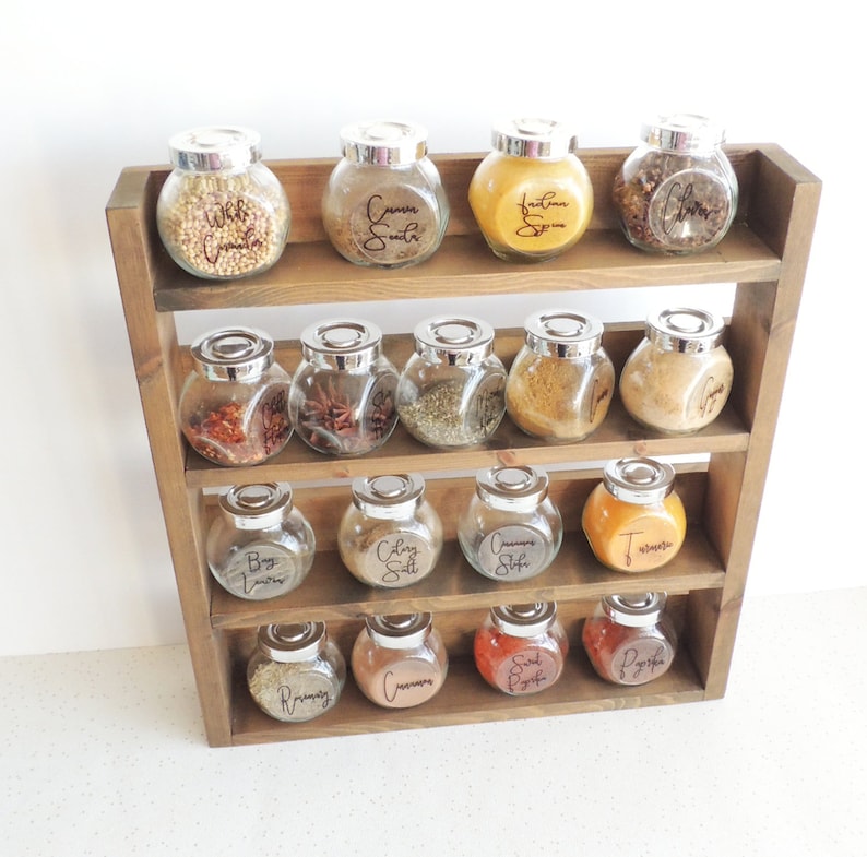 May include: A wooden spice rack with three shelves holds 15 glass jars with silver lids. Each jar has a handwritten label with the name of a spice, such as "Curry Powder", "Cinnamon Sticks", and "Salt".