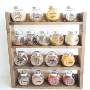 May include: A wooden spice rack with three shelves holds 15 glass jars with labels for various spices, including white cumin, cumin seeds, Italian spice, bay leaves, chili flakes, star anise, meat rub, ginger, turmeric, celery salt, cinnamon sticks, rosemary, cinnamon, sweet paprika, and paprika.