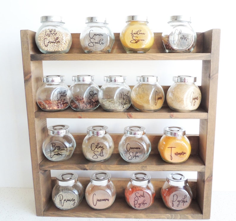 May include: A wooden spice rack with three shelves holds 15 glass jars with labels for various spices, including white cumin, cumin seeds, Italian spice, bay leaves, chili flakes, star anise, meat rub, ginger, turmeric, celery salt, cinnamon sticks, rosemary, cinnamon, sweet paprika, and paprika.