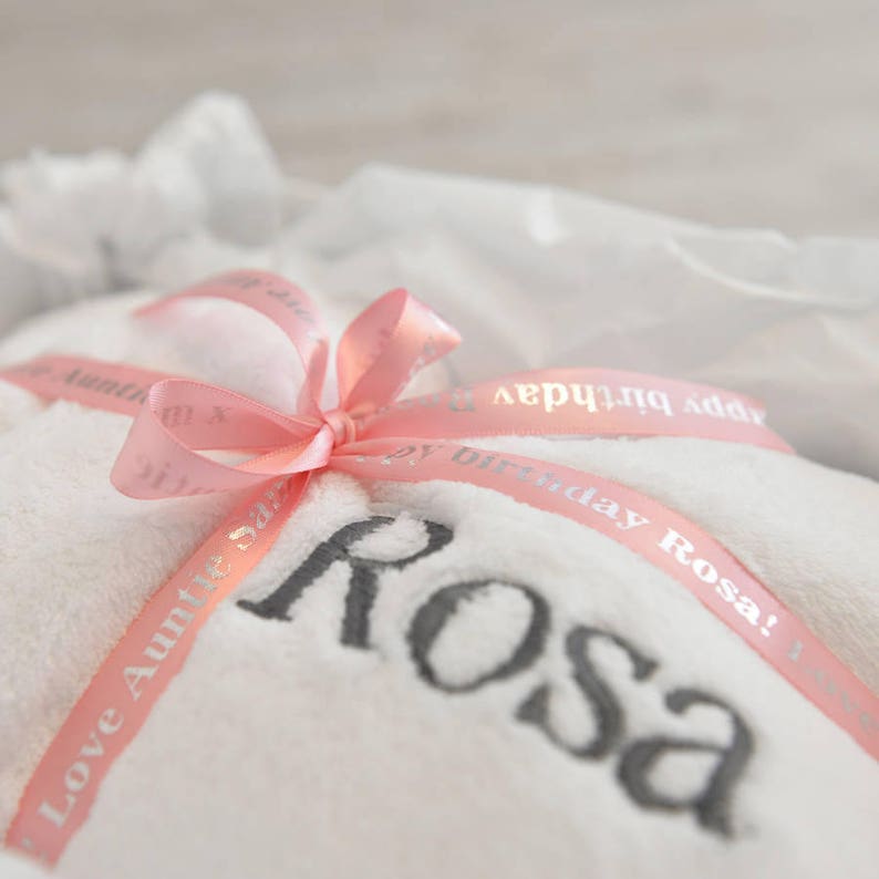 K&ouml;nnte beinhalten: Ein wei&szlig;es Handtuch mit dem Namen "Rosa" in grauen Buchstaben bestickt. Ein rosa Band mit dem Text "Happy Birthday Rosa!" und "Love Auntie Sasa" ist als Schleife um das Handtuch gebunden. Das Handtuch ist in wei&szlig;es Seidenpapier gewickelt.