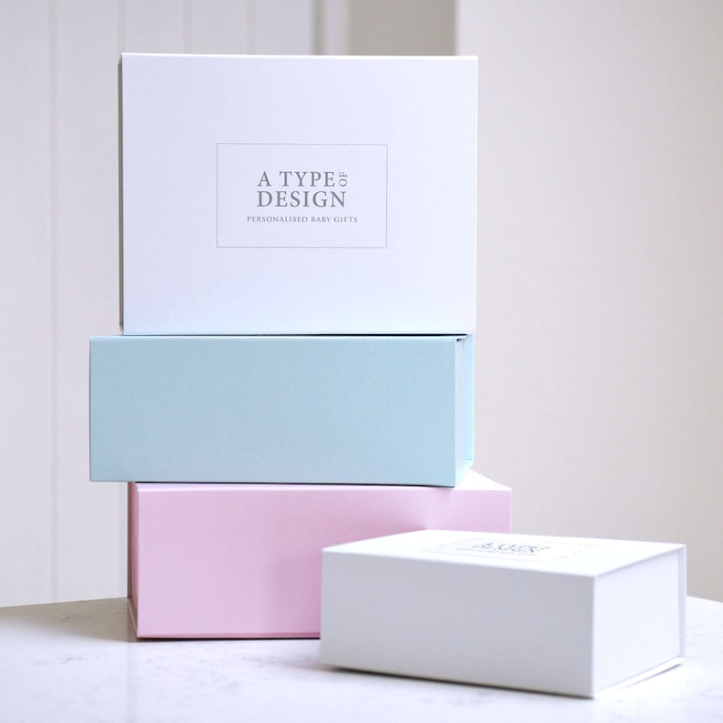 K&ouml;nnte beinhalten: Vier rechteckige Geschenkboxen in Pastellfarben: Wei&szlig;, Hellblau und Rosa. Die oberste Box ist wei&szlig; und tr&auml;gt den Text "A TYPE OF DESIGN PERSONALIZED BABY GIFTS". Die Boxen sind auf einer wei&szlig;en Oberfl&auml;che gestapelt.