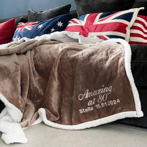 May include: A brown blanket with white sherpa trim, embroidered with "Amazing at 80 Stella 15.01.2024". The blanket is on a dark sofa with cushions displaying Union Jack and other flag designs.