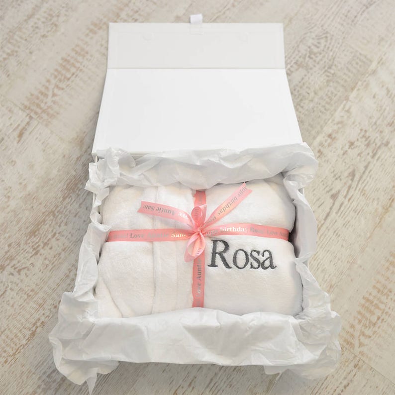 K&ouml;nnte beinhalten: Ein wei&szlig;er Bademantel mit dem Namen "Rosa" in Grau gestickt, pr&auml;sentiert in einer wei&szlig;en Geschenkbox. Der Bademantel ist mit einem rosa Band mit der Aufschrift "Happy Birthday" und "Love" gebunden. Die Box ist mit wei&szlig;em Seidenpapier ausgekleidet.