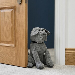 Op de afbeelding: Een grijze stoffen deurstopper in de vorm van een hond, met een zwarte kraag, geplaatst tussen een houten deur en een wit deurkozijn. De deurstopper heeft een visgraatpatroon en staat op een licht tapijt.