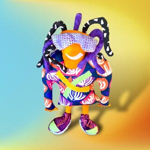 May include: A colorful handmade plush toy with a smiling orange face, purple hair, and black and white patterned clothing. The toy is wearing sunglasses and has a bright, playful expression.