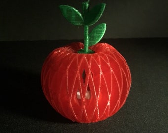 May include: A red, 3D-printed apple-shaped decorative object with a green stem and leaves. The apple has a lattice design with a small opening. The object is on a dark surface.