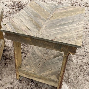 May include: A rustic wooden side table with a chevron pattern top and bottom shelf. The table is made of reclaimed wood and has a natural finish. The table is sitting on a sandy beach.