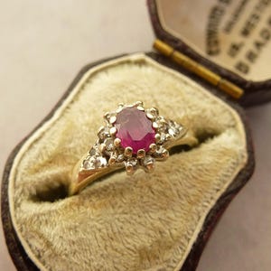 以下が含まれることがあります： リングボックスに入っている、ルビーの宝石を囲む小さなダイヤモンドがついた金の指輪