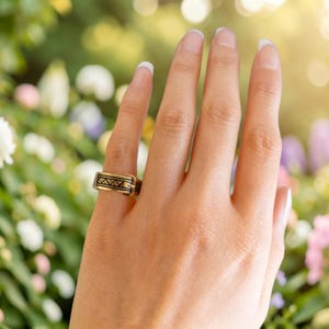 Puede incluir: Un anillo de tono dorado con forma de cintur&oacute;n en miniatura, que presenta un detalle de hebilla y un patr&oacute;n geom&eacute;trico negro grabado de tri&aacute;ngulos a lo largo de la banda. El anillo tiene un acabado pulido y se lleva en el dedo anular.