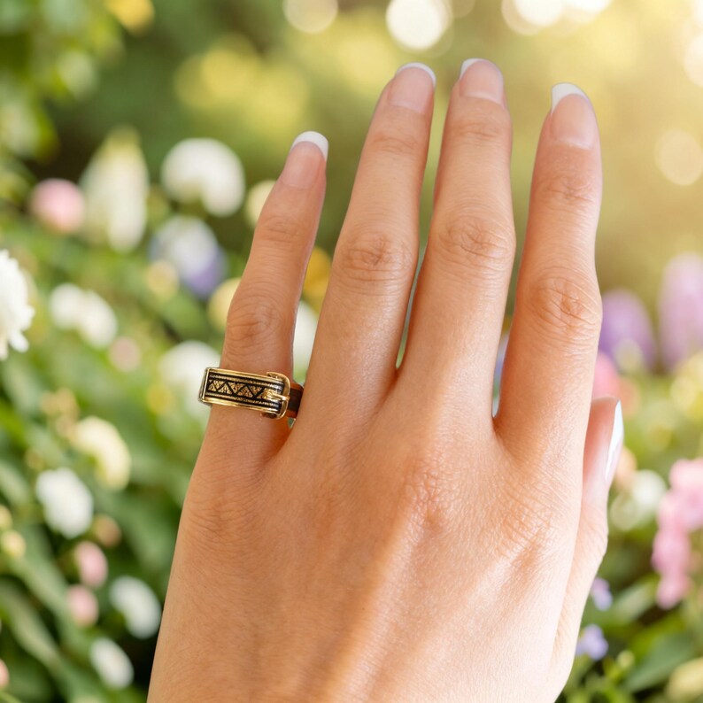 Puede incluir: Un anillo de tono dorado con forma de cintur&oacute;n en miniatura, que presenta un detalle de hebilla y un patr&oacute;n geom&eacute;trico negro grabado de tri&aacute;ngulos a lo largo de la banda. El anillo tiene un acabado pulido y se lleva en el dedo anular.